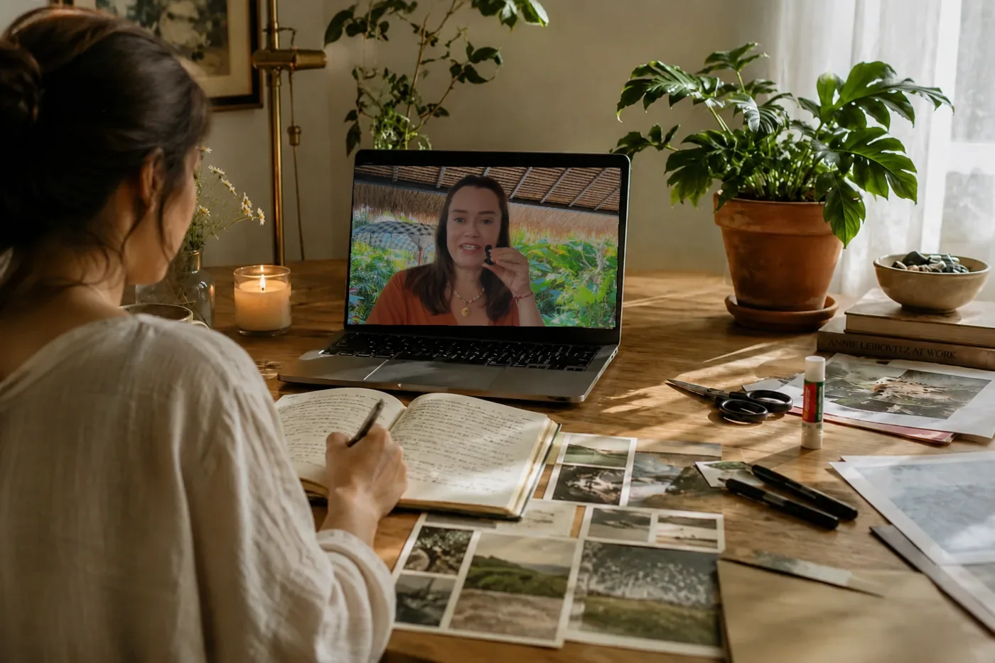 A woman at her desk writing in a notebook beside collage clippings, an online workshop on her laptop