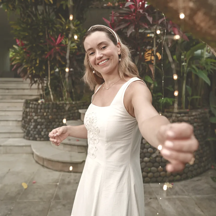 A woman in a white dress smiling with a sparkler in a garden at dusk