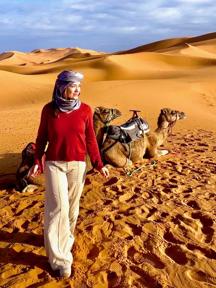 Ana walking across desert dunes, camels resting behind her
