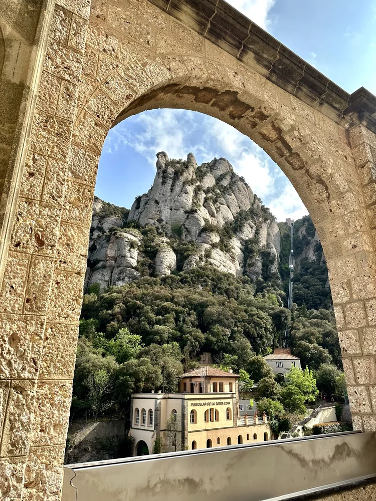 A stone arch framing jagged mountains and an old building in the trees